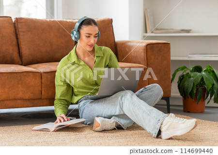 A young woman is seated on the floor of a well-lit living room with her back against a couch, focused on a laptop placed on her lap. She wears casual clothing and headphones, and has a notebook A young woman is seated on the floor of a well-lit living room with her back against a couch, focused on a laptop placed on her lap. She wears casual clothing and headphones, and has a notebook 114699440