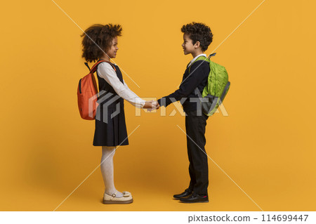African American young boy and girl, both dressed in school uniforms with backpacks, are standing against a vibrant yellow backdrop, holding hands African American young boy and girl, both dressed in school uniforms with backpacks, are standing against a vibrant yellow backdrop, holding hands 114699447