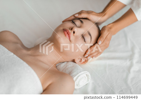 A young blonde woman enjoys a relaxing facial massage at a spa, embodying serenity and tranquility in a soothing environment 114699449