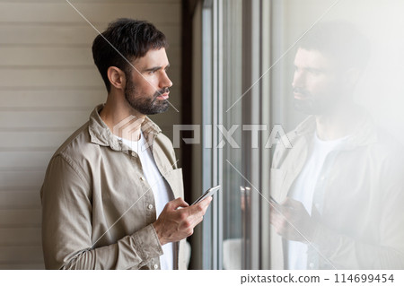 A man is standing in front of a window, looking at his cell phone. He appears focused on the screen, with natural light coming through the window. 114699454
