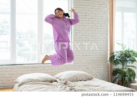 A middle eastern woman is energetically jumping in the air on top of a neatly made bed in a bright room. She appears to be mid-jump with her arms stretched out and a joyful expression on her face. A middle eastern woman is energetically jumping in the air on top of a neatly made bed in a bright room. She appears to be mid-jump with her arms stretched out and a joyful expression on her face. 114699455