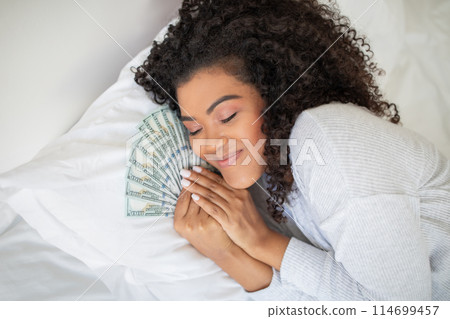 A Hispanic woman is seen laying on a bed with a large amount of money spread out around her. She appears to be counting or looking at the money. 114699457