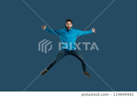 A man wearing a blue shirt is captured mid-air as he jumps energetically. His body is stretched upwards with his arms raised, showcasing a moment of dynamic movement. A man wearing a blue shirt is captured mid-air as he jumps energetically. His body is stretched upwards with his arms raised, showcasing a moment of dynamic movement. 114699482