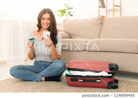 A young woman is seated comfortably on the floor with her legs crossed in a casual setting. She is smiling while engaged with her smartphone, holding credit card. Beside her, an open suitcase 114699495