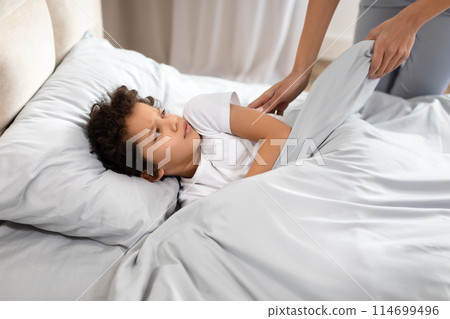 African American young child rests on a bed with a look of anticipation, watching as a parent lovingly adjusts the blanket over them 114699496