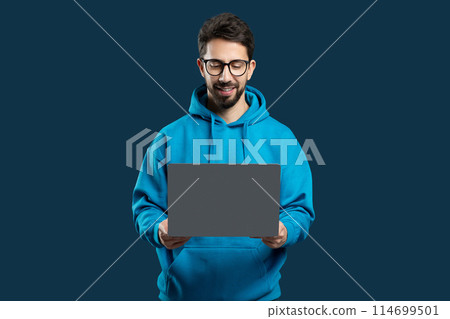 A man wearing a blue hoodie is standing while holding a laptop in his hands. He appears focused on using the device and possibly working or browsing the internet. A man wearing a blue hoodie is standing while holding a laptop in his hands. He appears focused on using the device and possibly working or browsing the internet. 114699501