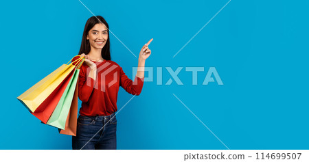 A cheerful young woman dressed in a casual red top and blue jeans stands against a solid blue backdrop, holding colorful shopping bags in one hand while pointing at copy space A cheerful young woman dressed in a casual red top and blue jeans stands against a solid blue backdrop, holding colorful shopping bags in one hand while pointing at copy space 114699507