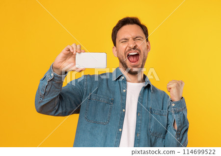 A man in a denim shirt is exuberantly celebrating, eyes closed, with a clenched fist and a wide-open mouth as if cheering or shouting. He holds a smartphone in his other hand 114699522