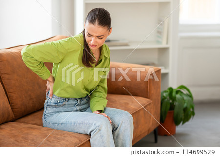 A woman is visibly in discomfort, pressing on her lower back with one hand while sitting on the edge of a brown couch in a well-lit living room, suggesting she is suffering from back pain. 114699529
