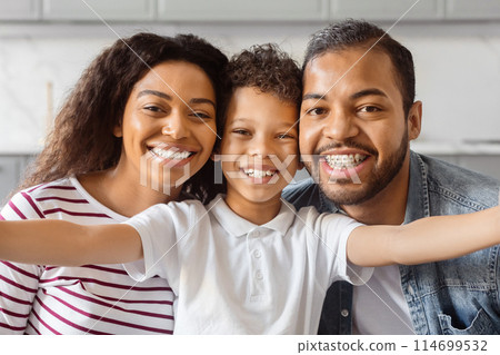 African American man, woman, and child are smiling and posing together while taking a selfie. They are holding a smartphone at arms length and capturing a candid moment of joy and togetherness. 114699532
