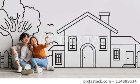 A young, smiling asian couple is casually seated on the floor, leaning against a white wall with a line drawing of a house and a tree 114699534