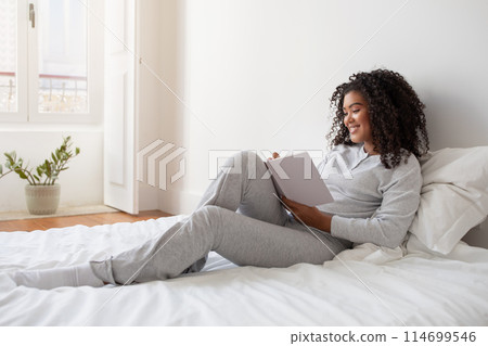 A Hispanic woman in casual clothing sitting on a neatly made bed, engrossed in writing in diary. The room appears cozy with soft lighting, and there are a few pillows scattered on the bed. 114699546