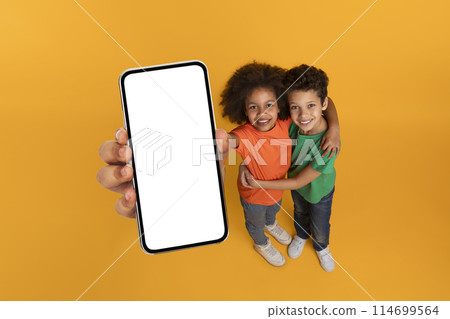 African American kids holding up a cell phone with a blank white screen, showing no content or images, mockup copy space, yellow studio background African American kids holding up a cell phone with a blank white screen, showing no content or images, mockup copy space, yellow studio background 114699564