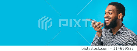Happy young black man using voice assistant on smartphone, isolated over blue studio wall, copy space, web-banner 114699582