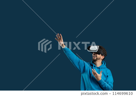 A man wearing a blue hoodie is immersed in a virtual reality experience while wearing a headset. He is interacting with a digital environment as he explores and engages with the virtual world A man wearing a blue hoodie is immersed in a virtual reality experience while wearing a headset. He is interacting with a digital environment as he explores and engages with the virtual world 114699610