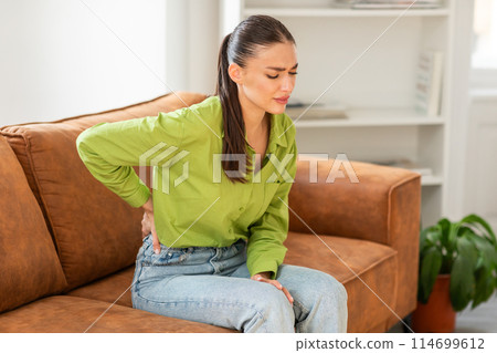 Young woman, dressed in green blouse and blue jeans, sits on a brown leather sofa with a pained expression, clutching her lower back with one hand, indicating discomfort or a potential back problem. 114699612
