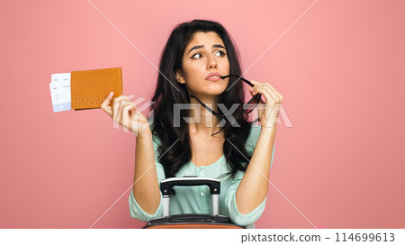 A young middle eastern woman traveler holds her passport and sunglasses, looking up thoughtfully while leaning on the handle of her suitcase. She seems to be pondering her next travel destination 114699613