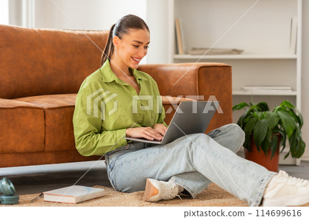 A woman is sitting on the floor, focused on using a laptop in front of her. She appears engaged in her work, with a serious expression on her face. 114699616