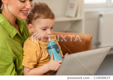 A woman is sitting while holding a child in her arms, multitasking by using a laptop. Mother and son watching cartoon together, home interior, copy space A woman is sitting while holding a child in her arms, multitasking by using a laptop. Mother and son watching cartoon together, home interior, copy space 114699637