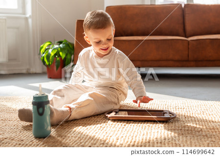 A young boy is seated on the floor, engrossed in playing with a tablet device. His focus is on the screen as he interacts with the digital content 114699642