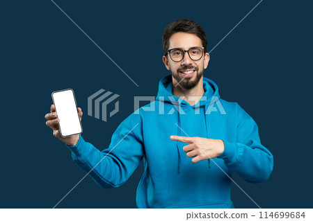 A cheerful young man with a beard and glasses is wearing a blue hoodie and presenting a smartphone with its screen facing forward towards the viewer. He is pointing at the screen 114699684