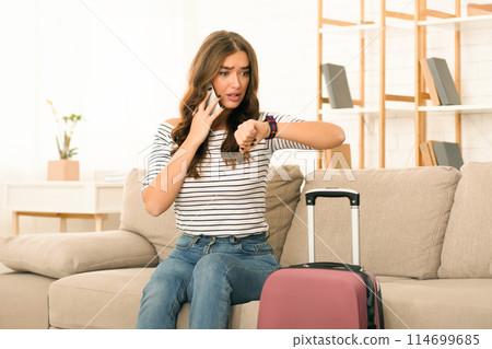 A concerned young woman sits on the edge of a couch, gazing at her watch with a look of anxiety on her face. She is preparing to depart, evident by a suitcase beside her A concerned young woman sits on the edge of a couch, gazing at her watch with a look of anxiety on her face. She is preparing to depart, evident by a suitcase beside her 114699685