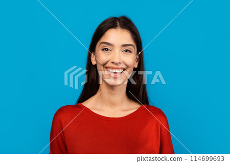 A woman wearing a red top smiles directly at the camera, radiating a sense of joy and confidence in her expression. She appears relaxed and at ease, exuding positivity. 114699693