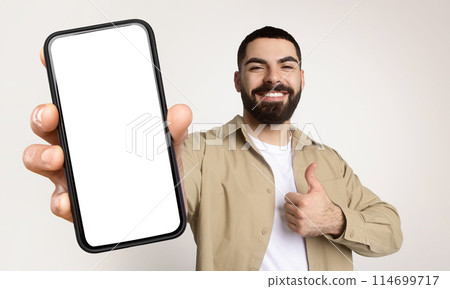 Millennial man is holding a smart phone with white blank screen in his hand, showing thumb up, isolated on beige studio background Millennial man is holding a smart phone with white blank screen in his hand, showing thumb up, isolated on beige studio background 114699717