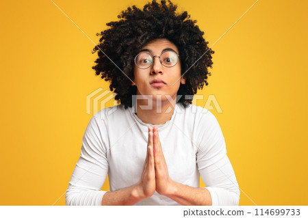 Young black man showing clasped hands in pleading gesture, pretty please on orange studio background 114699733