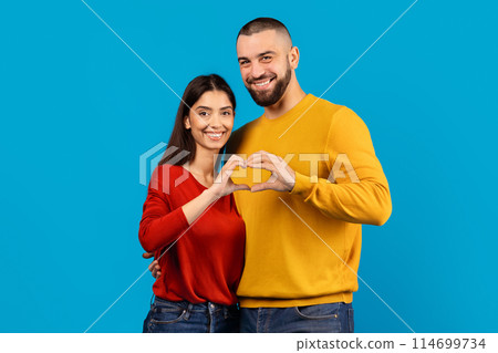 A cheerful man and woman stand close together against a vivid blue backdrop, their fingers intertwined to form a heart shape, symbolizing love and connection A cheerful man and woman stand close together against a vivid blue backdrop, their fingers intertwined to form a heart shape, symbolizing love and connection 114699734