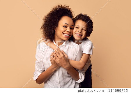A joyful African American mother holds her son close in a tender hug, both wearing white shirts, showcasing the affectionate bond between parent and child 114699735