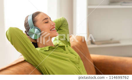 A woman is seated on a couch, wearing headphones. She appears to be listening to music or watching a video, immersed in the content playing through the headphones, copy space 114699762