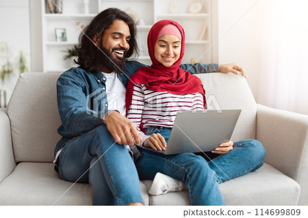 A young eastern couple using a laptop together in a sunlit living room, reflecting a cozy, connected atmosphere at home. 114699809