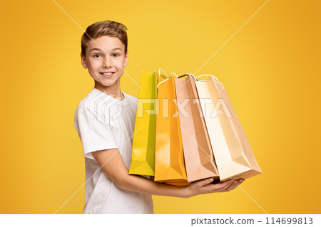Little shopper. Happy teen boy holding lots of colorful paper shopping bags in hands, orange studio background 114699813