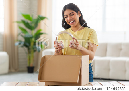 A young middle eastern woman stands in her sunlit living room, filled with excitement and a bright smile as she unpacks a cardboard box on a table, holding headphones 114699831