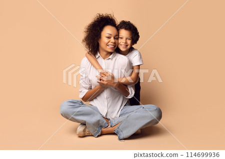 African American woman is seated on the ground, next to her is a child. Mother appears to be interacting with her son, possibly engaged in conversation or playing together. 114699936