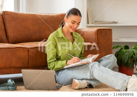 A woman is sitting on the floor, using a laptop and a notebook. She appears focused as she works on her tasks. The laptop screen emits a soft glow in the room. A woman is sitting on the floor, using a laptop and a notebook. She appears focused as she works on her tasks. The laptop screen emits a soft glow in the room. 114699956