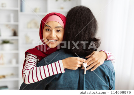 A young eastern lady wearing a red hijab is hugging her husband from behind, holding key from their new house 114699979