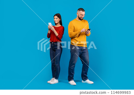 A man in a yellow sweater and jeans and a woman in a casual red top with dark denim jeans stand side by side, each absorbed in their own smartphone, blue background A man in a yellow sweater and jeans and a woman in a casual red top with dark denim jeans stand side by side, each absorbed in their own smartphone, blue background 114699983