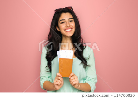 A cheerful young middle eastern woman stands against a vibrant pink backdrop, clutching a passport and a boarding pass, suggesting she is prepared and excited for an upcoming journey 114700020