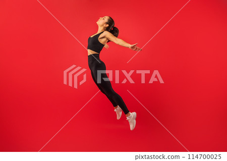 A woman in black sports bra and leggings is mid-air, jumping energetically. She is captured in a moment of athleticism and motion, showcasing her strength and agility. 114700025