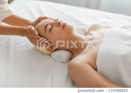 A woman lying down with her eyes closed, receiving a gentle massage on her face from a professional aesthetician using soothing motions. 114700033