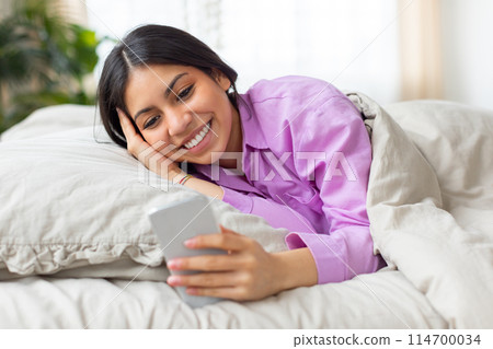 A cheerful young middle eastern woman lies comfortably in bed, wearing a pink shirt, as she enjoys browsing on her smartphone in the softly lit calmness of the morning 114700034