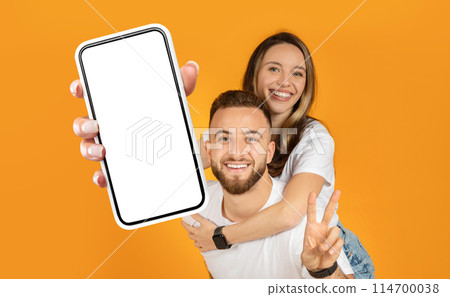 Cheerful young couple standing together, both holding up a cell phone towards the camera. They are looking at the phones screen, possibly taking a selfie or video call. 114700038