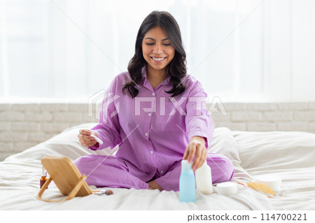 A cheerful young middle eastern woman wearing purple pajamas is sitting cross-legged on her bed engaged in her morning skincare routine 114700221