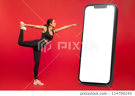 Flexible young woman in black sportswear exercising next to huge phone with white blank screen, on red studio background, practicing yoga or pilates, full length 114700243