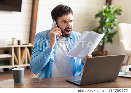 An adult man in a casual blue shirt is seated at a wooden desk in a well-lit home office. He appears agitated while talking on a mobile phone and holding a piece of paper An adult man in a casual blue shirt is seated at a wooden desk in a well-lit home office. He appears agitated while talking on a mobile phone and holding a piece of paper 114700275