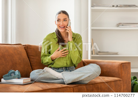 A woman is seated on a couch, engaged in a conversation on her cell phone. She appears focused on the call, with a concentrated expression on her face. A woman is seated on a couch, engaged in a conversation on her cell phone. She appears focused on the call, with a concentrated expression on her face. 114700277
