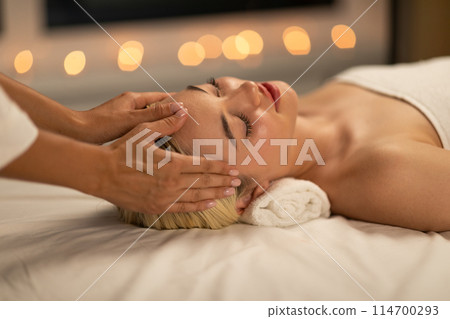 In a tranquil spa environment, a young woman with blonde hair experiences a soothing facial massage, radiating relaxation and peace. 114700293