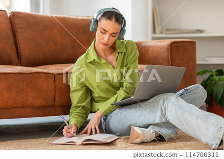 A focused young woman is sitting on the floor with her back against a couch in a well-lit room, listening to music on headphones while writing in a notebook, with a laptop open in front of her. A focused young woman is sitting on the floor with her back against a couch in a well-lit room, listening to music on headphones while writing in a notebook, with a laptop open in front of her. 114700311